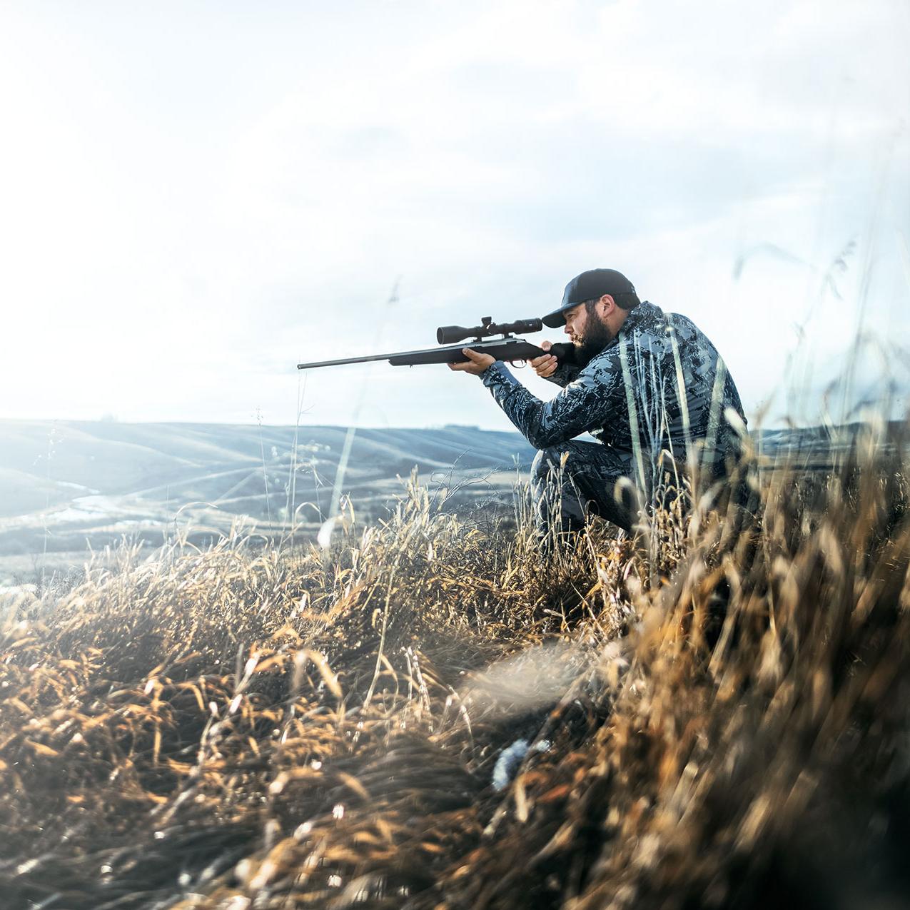En mann kneler i gresset, med et vidstrakt, bølgende landskap i bakgrunnen, og sikter med riflen sin.