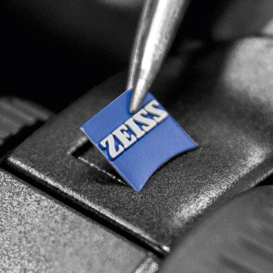 Et ZEISS-logoinnlegg plasseres med en tang på en svart metalloverflate.
