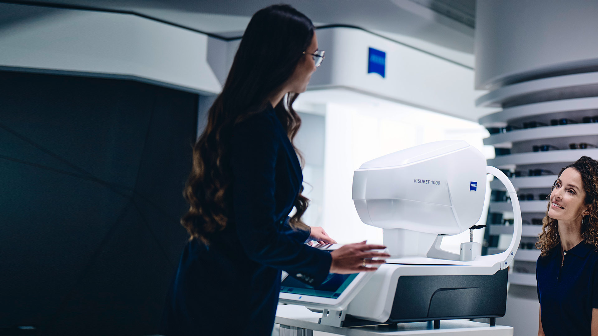 En kvinne sitter foran ZEISS VISUREF 1000 og ser mot en kvinnelig optiker.