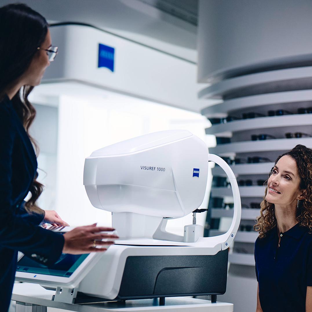 En kvinne sitter foran ZEISS VISUREF 1000 og ser mot en kvinnelig optiker.