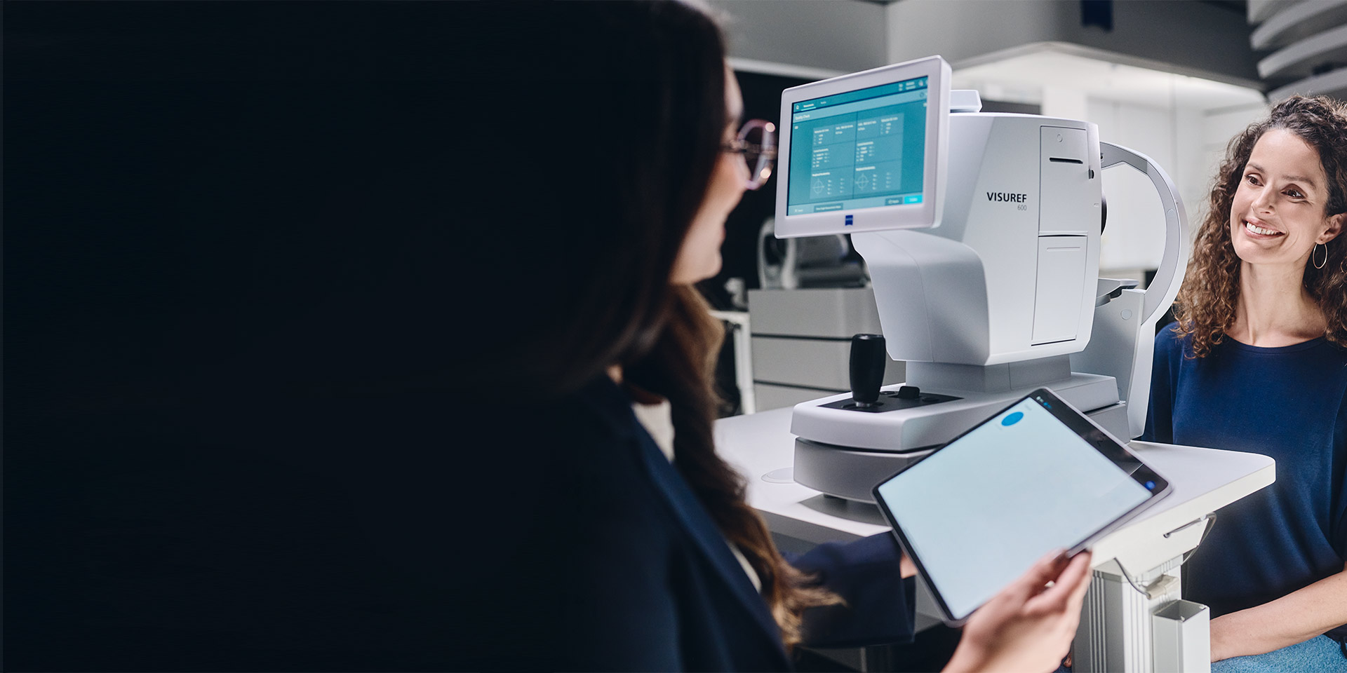 En kvinnelig optiker ser på nettbrettet sitt mens hun sitter foran ZEISS VISUREF 600 og snakker med kunden.