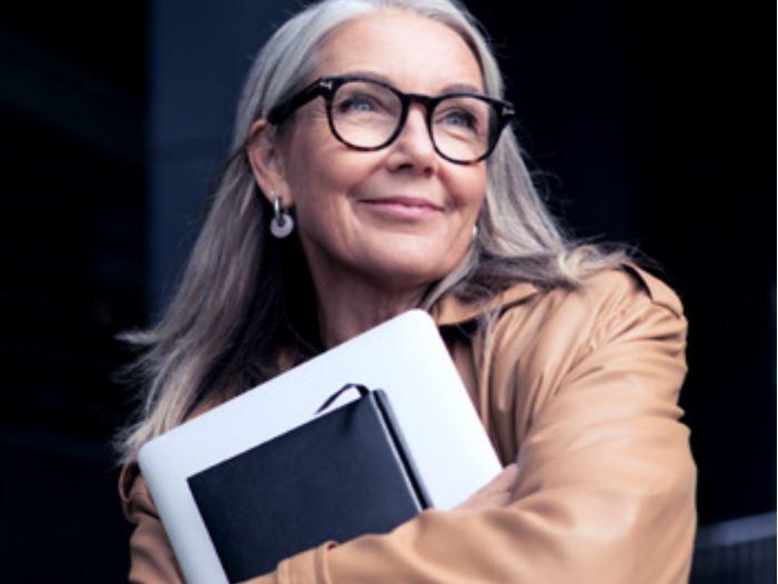 Et bilde av en eldre kvinne med briller som smiler mens hun bærer en bærbar datamaskin.