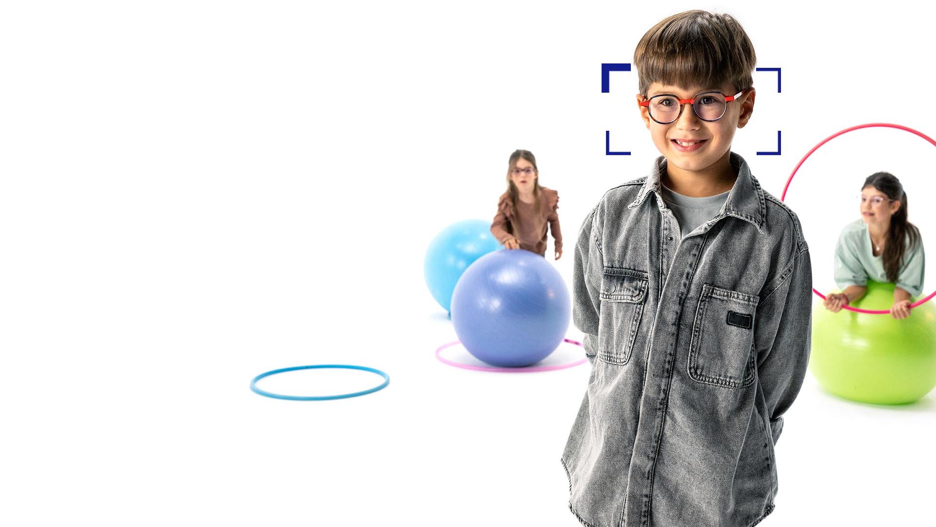 Brunhåret gutt med runde briller med ZEISS MyoCare-brilleglass står i forgrunnen og smiler til kameraet. I bakgrunnen er to jenter med ZEISS MyoCare-brilleglass som leker med bøyler og gymnastikkballer.