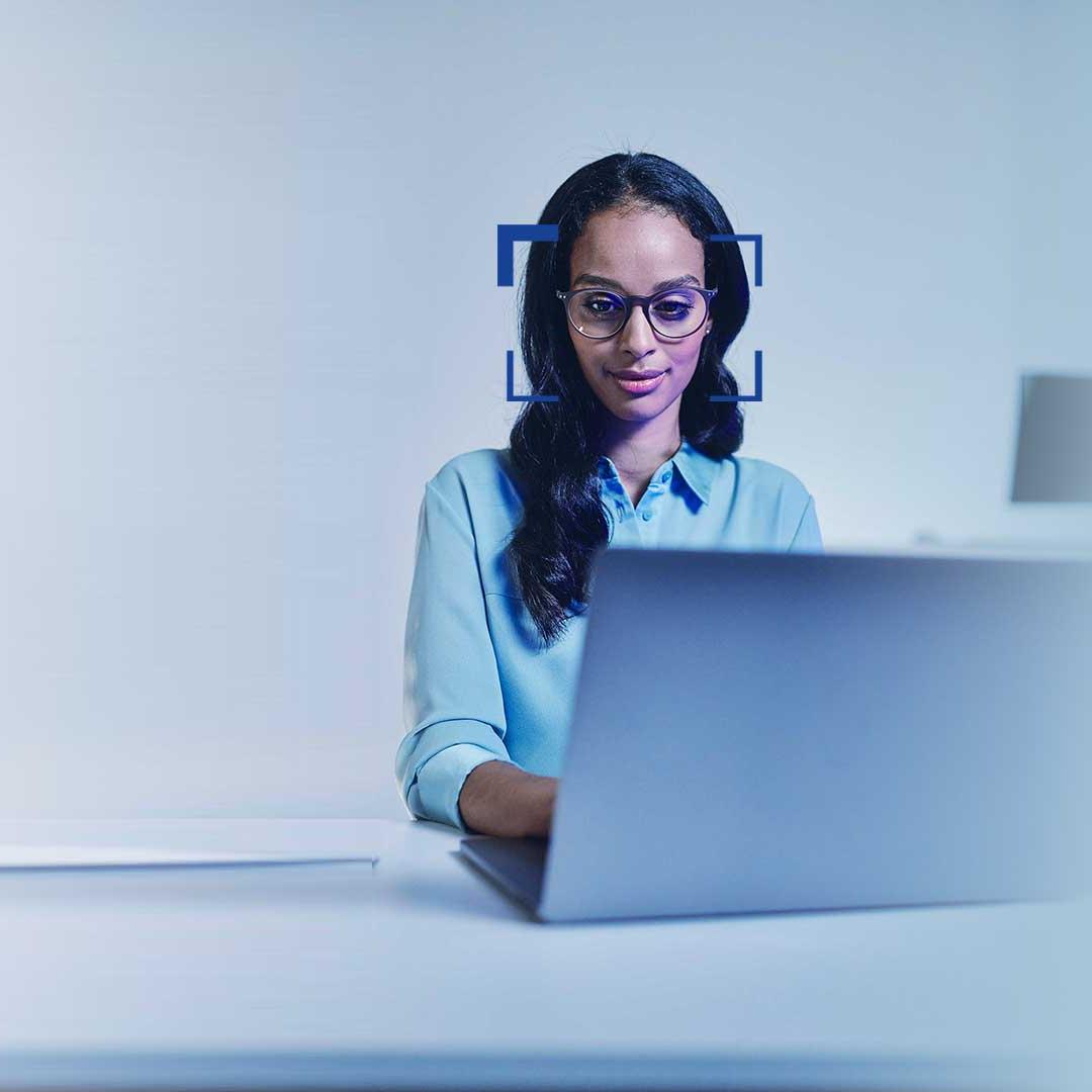 Kvinne med svart hår og briller ser smilende inn i en bærbar datamaskin