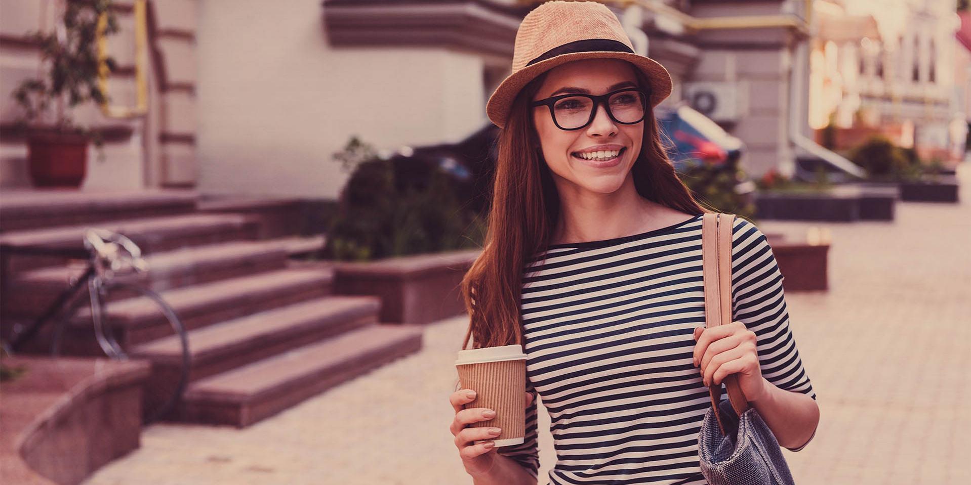 En ung kvinne med caps og briller bærer et kaffekrus (høyre side) og en bag på venstre skulder