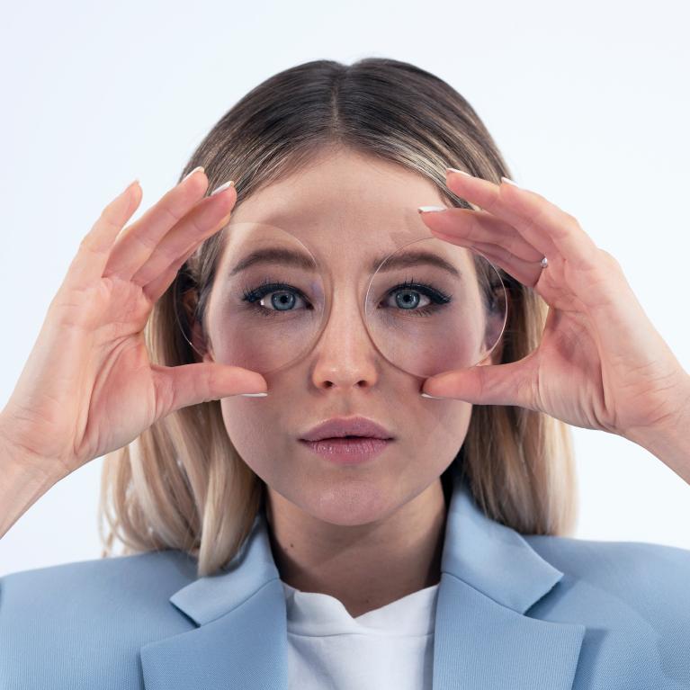 En ung blond kvinne holder glass foran øynene for å vise et godt utseende uten noen morsomme øyeeffekter takket være ZEISS ClearView enstyrkeglass.