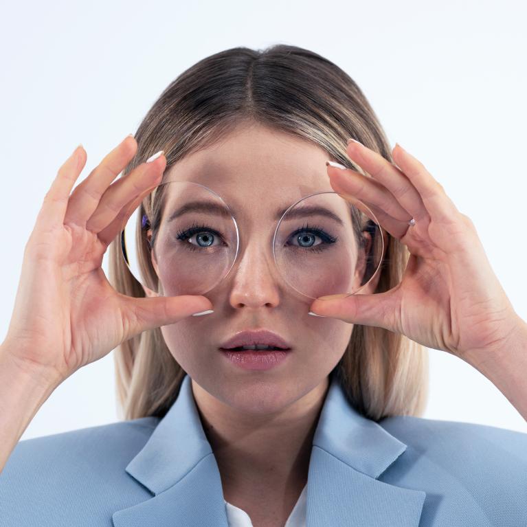 En ung blond kvinne holder glass foran øynene for å vise små-øyneeffekten forårsaket av tykke minusbriller.