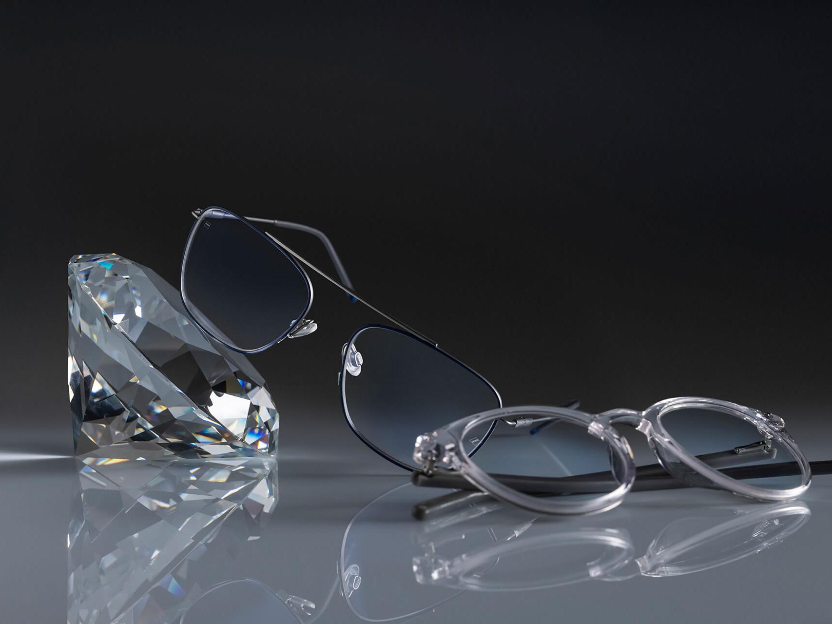 To par briller med ZEISS-glass som har det hardeste ZEISS-belegget noensinne - DuraVision® Platinum. Den ene brillen lener seg mot en krystall, den andre ligger flatt på bakken. Begge har klare glass uten blåaktig refleksjon.