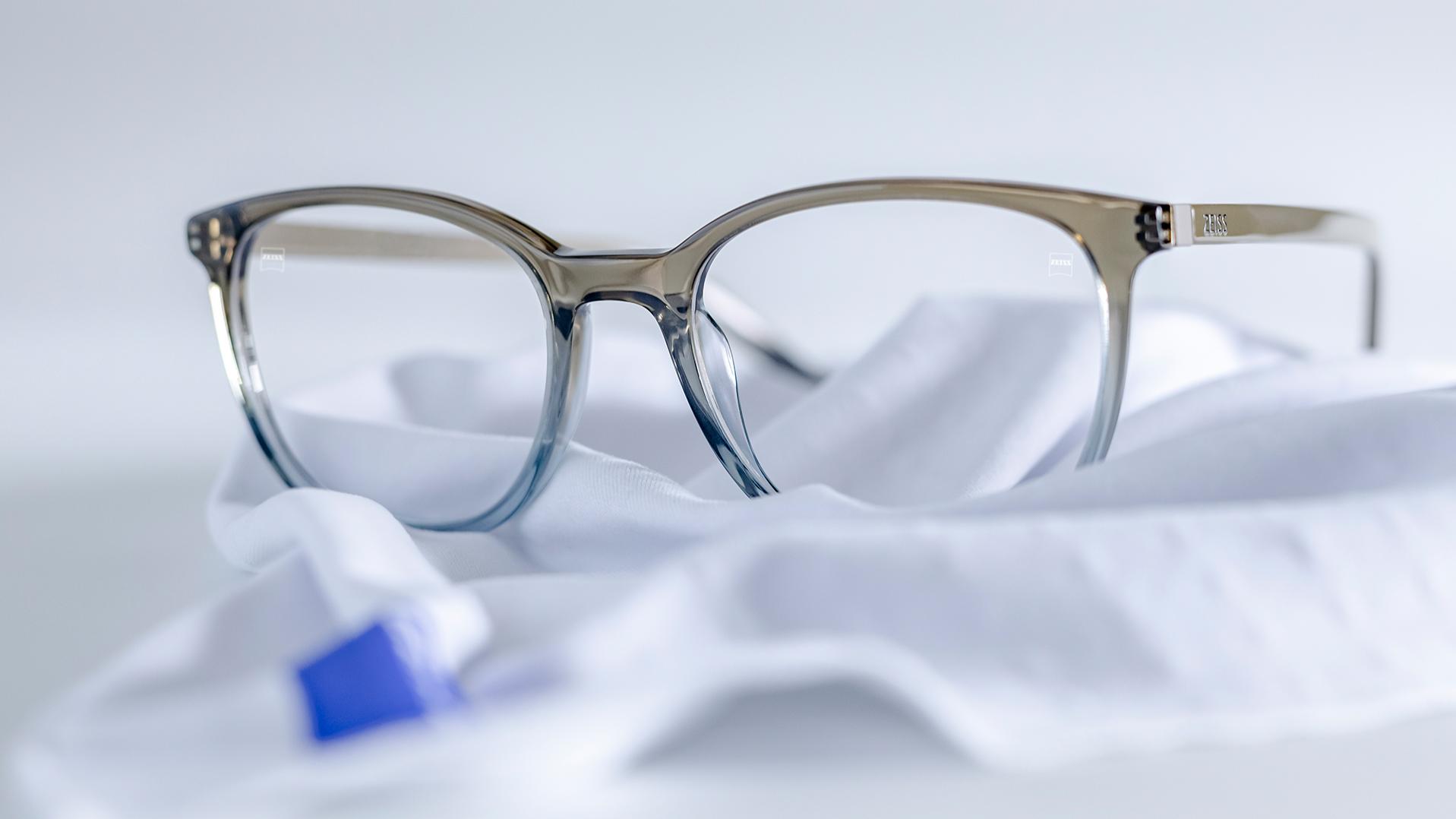 Et par briller med gråblå innfatning og ZEISS-glass med DuraVision®-belegg ligger på en hvit mikrofiberklut.