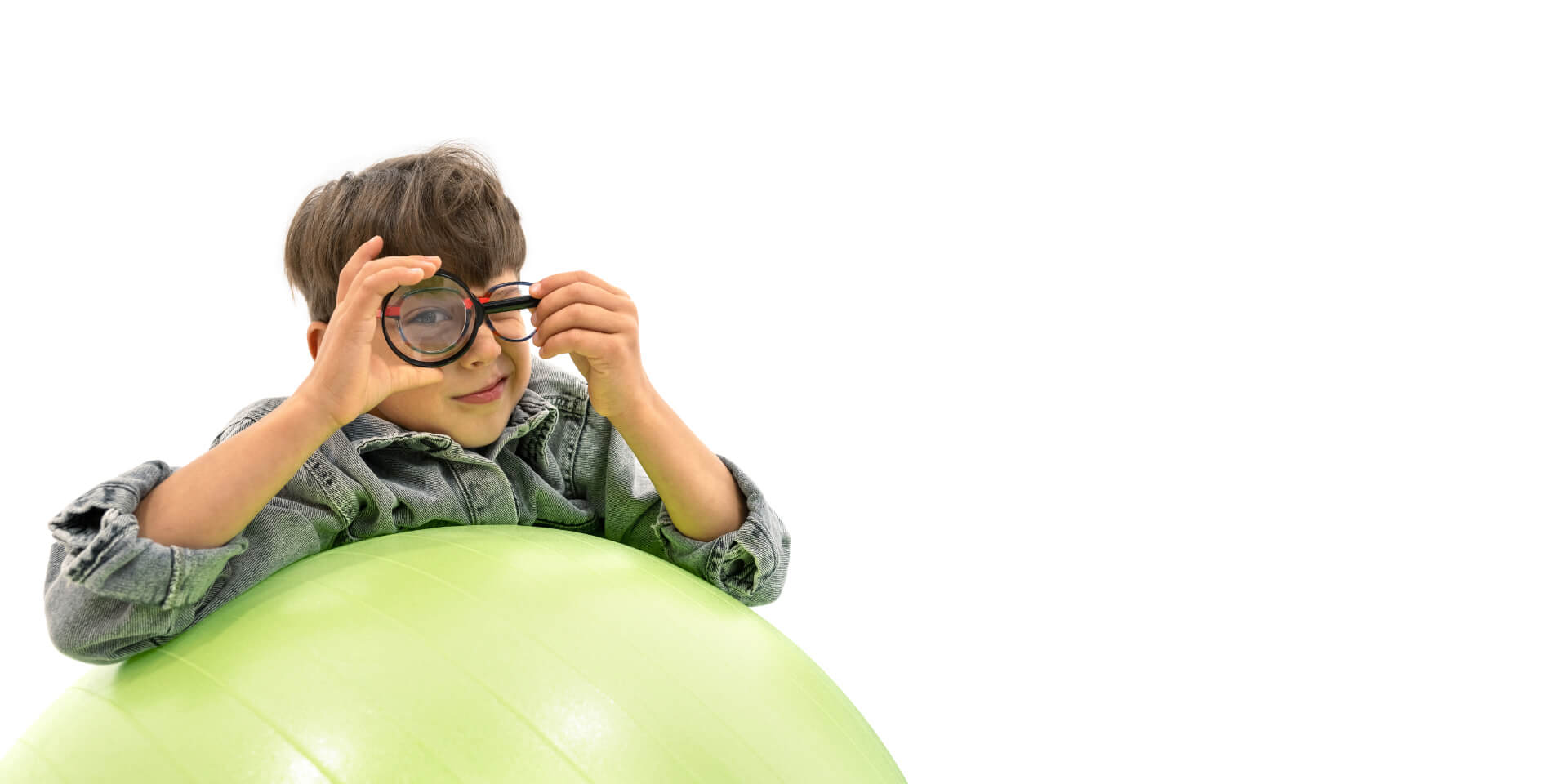 En gutt med ZEISS-glass for behandling av nærsynthet som lener seg på en gymnastikkball og holder et forstørrelsesglass foran det ene øyet.