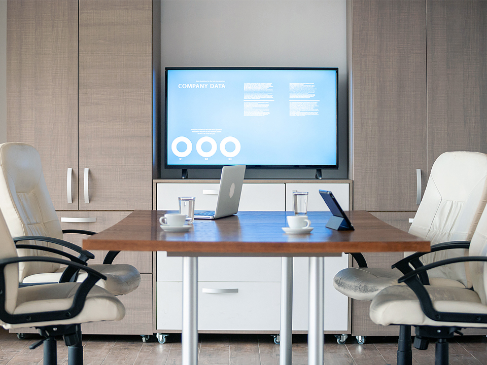Utsikt fra skrivebordet på et lederkontor. En skjerm er montert på en vegg noen meter unna. Det er et lite møtebord med kontorstoler og bærbare datamaskiner på bordet.