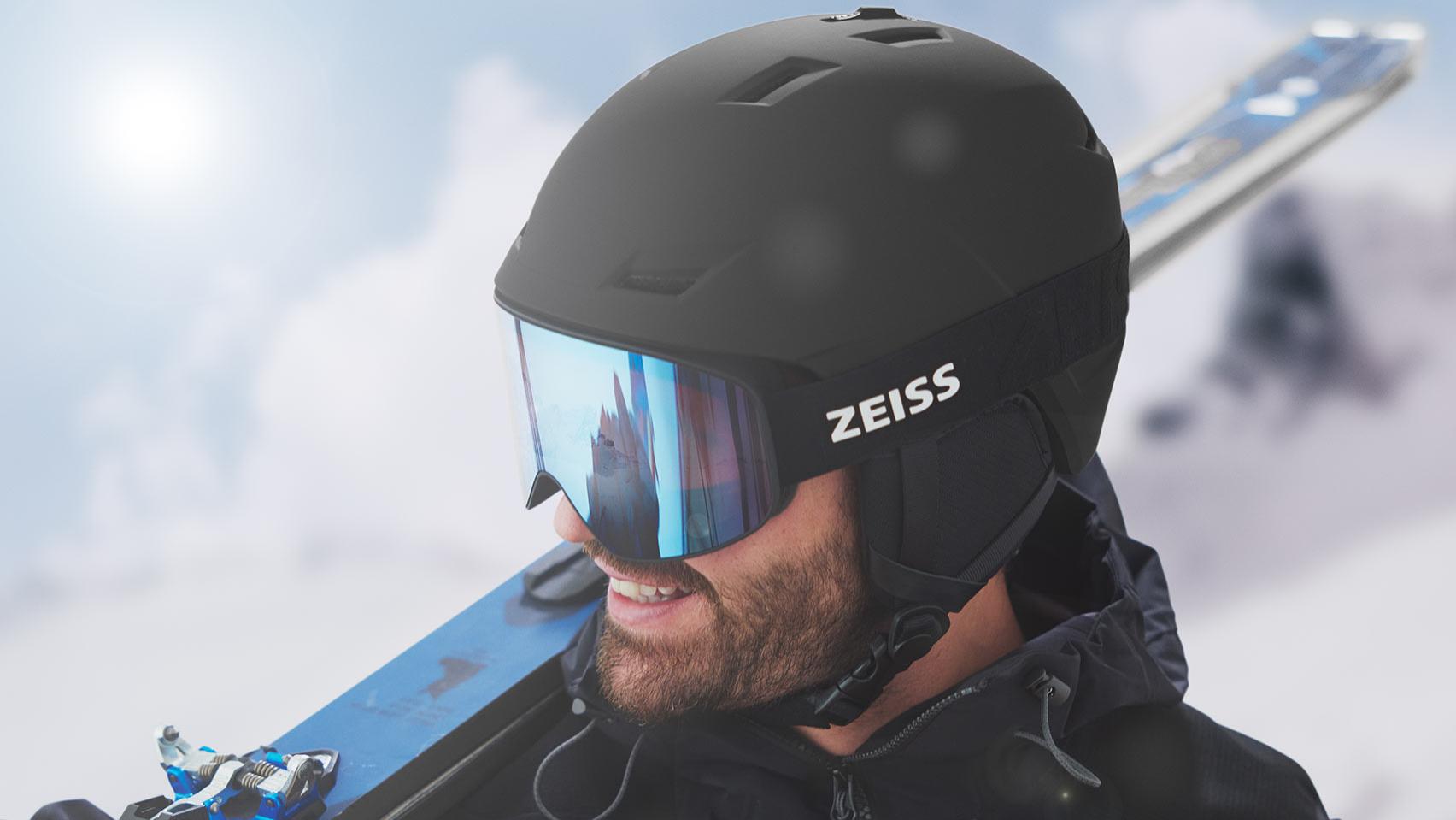 En kvinne bruker en svart hjelm med blå ZEISS skibriller med speilglass og holder et par ski over høyre skulder.