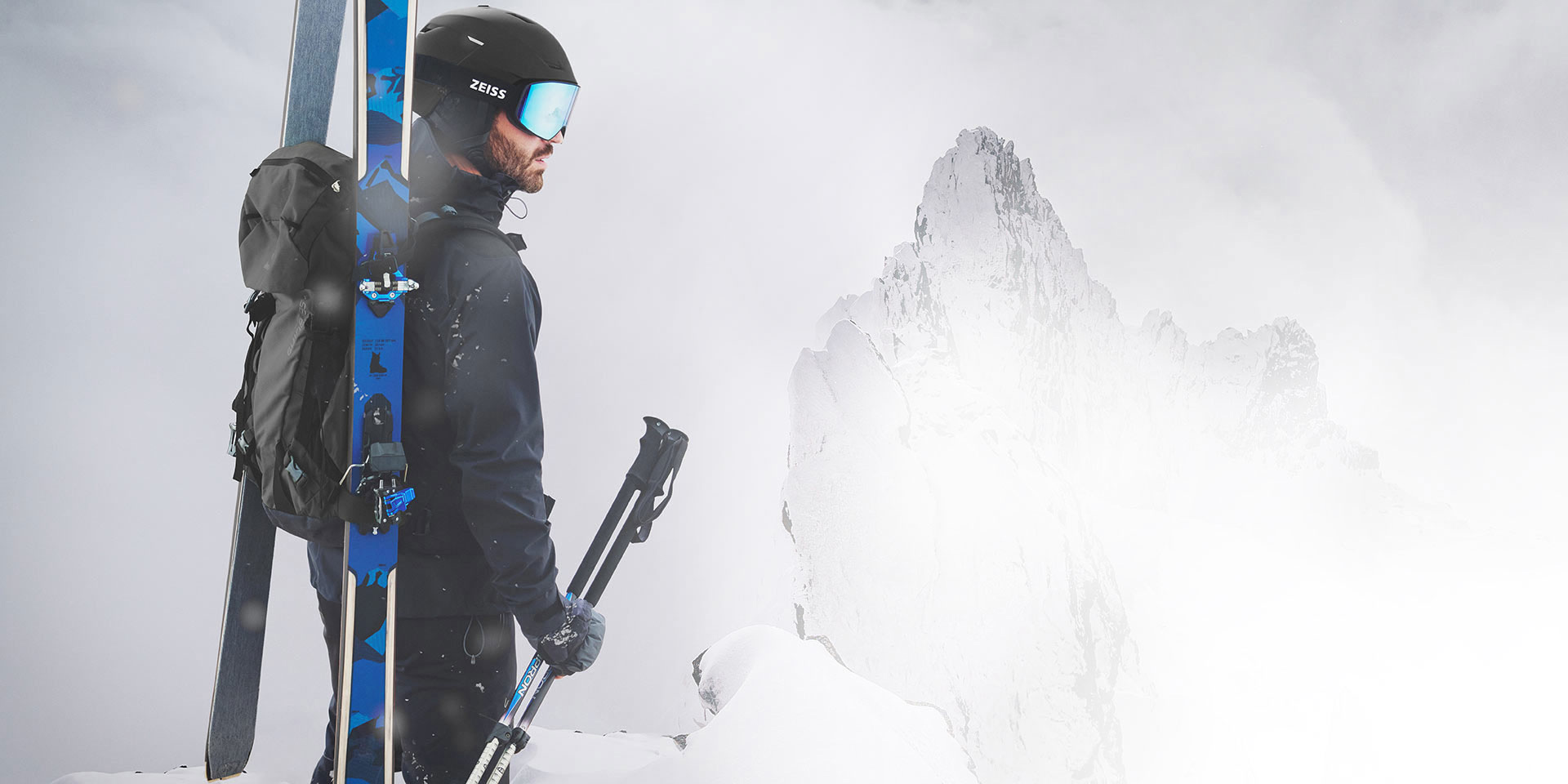 En mann bruker en svart hjelm med blå speilede ZEISS skibriller. Han har ski og en ryggsekk på ryggen, samtidig som han holder skistavene sine i høyre hånd. Han står på en fjelltopp.