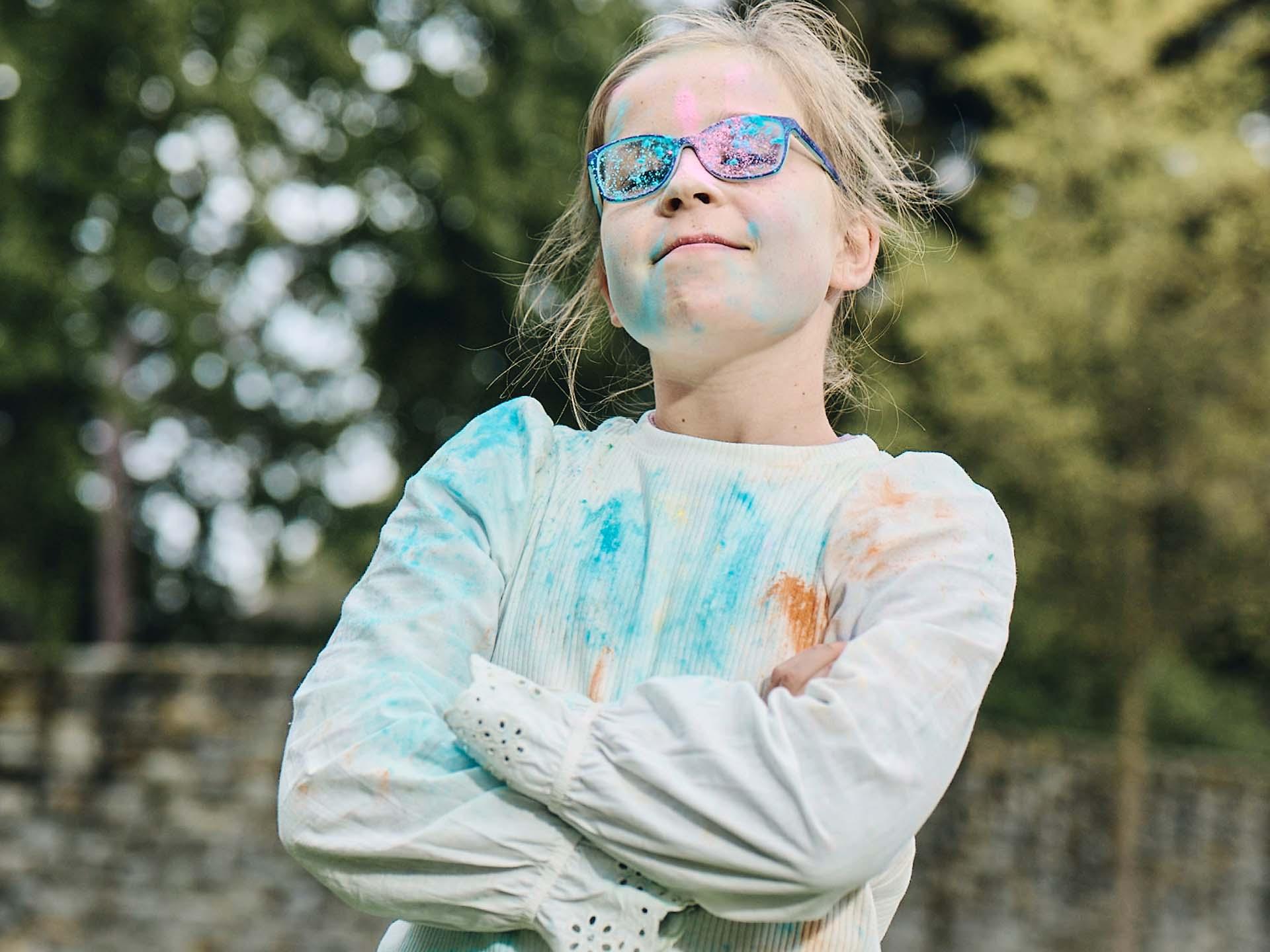 En jente med korslagte armer og skitne briller etter å ha lekt med farget pulver ser modig ut og smiler.
