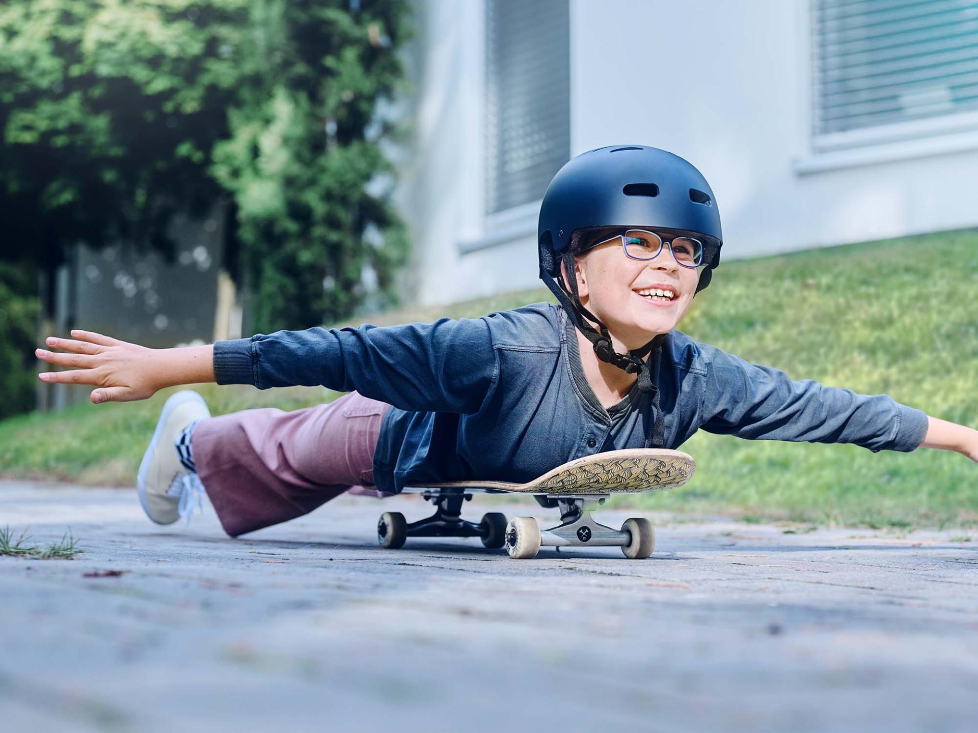 En jente med hjelm og briller ruller liggende på et skateboard nedover veien og strekker armene ut.